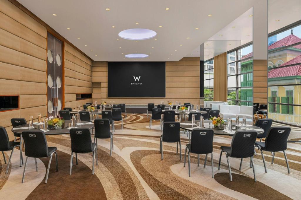 A daylight meeting room with round tables, black chairs, a large screen and tall windows overlooking nearby buildings.