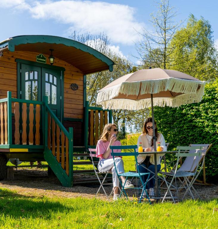 two women sitting at a table under an umbrella in front of a cabin at Les Roulottes & Cottages du Moulin in Chenillé-Changé