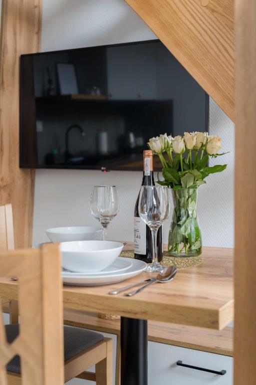 Small dining table set with plates, wine bottle, two glasses, vase of white roses, wall-mounted TV.