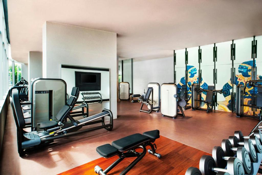A bright hotel gym with strength machines, a bench, dumbbells, and a television.