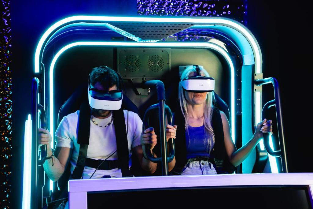 Pair wearing VR headsets seated in neon-lit simulator pod, gripping safety handles.