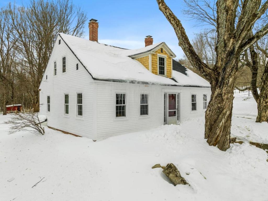 a white house in the snow with a tree at Mini Cows, Trails & Cozy Porch in Gilmanton