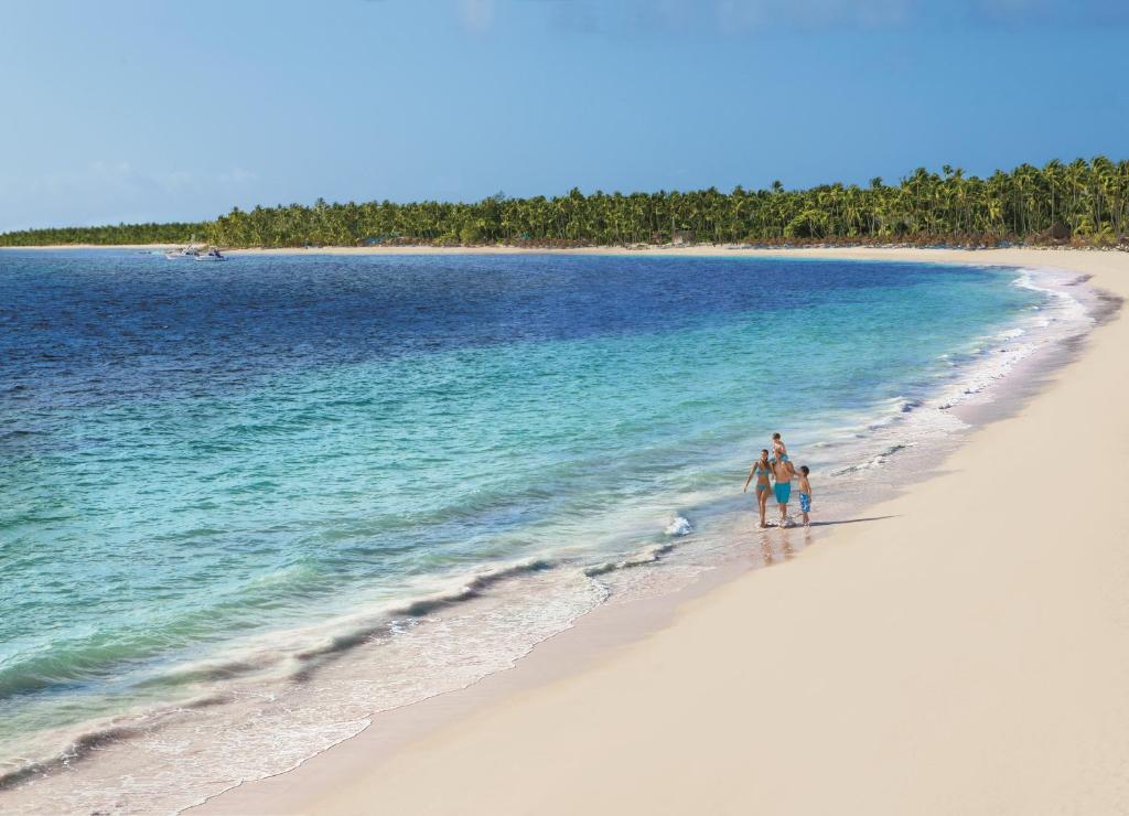 Sandy beach with turquoise ocean water, guests wading, and palm-lined shore in distance.