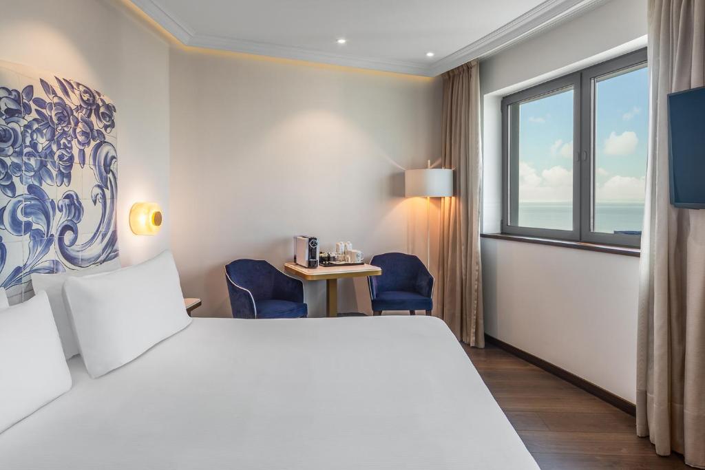A bright room with a large white bed, compact sitting area and a broad window.