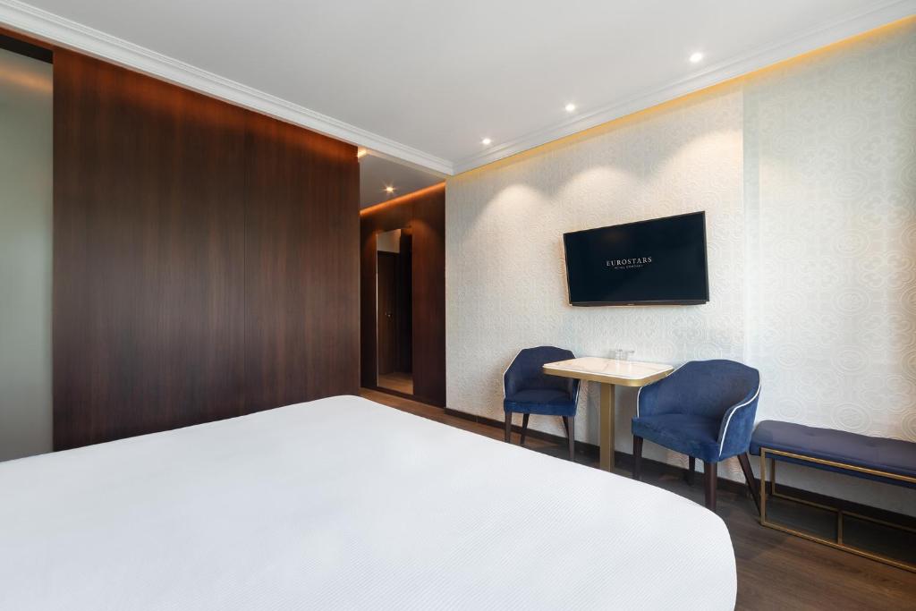 A room view showing the bed, a two-chair table, television and dark wood storage wall.