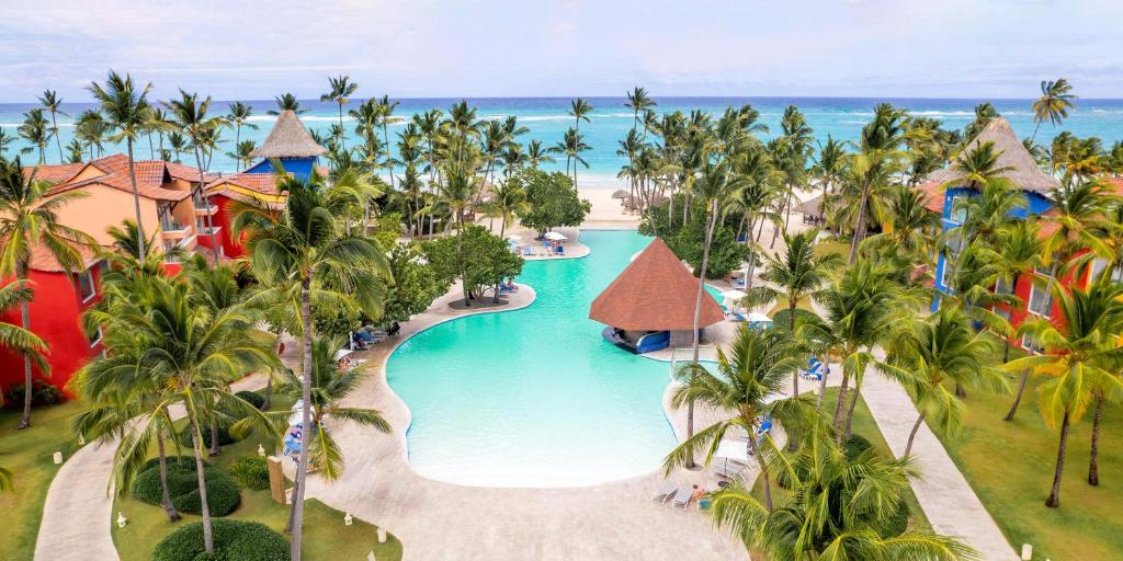 Resort aerial view showing curved pool with thatched-roof structures and palm trees near beach.