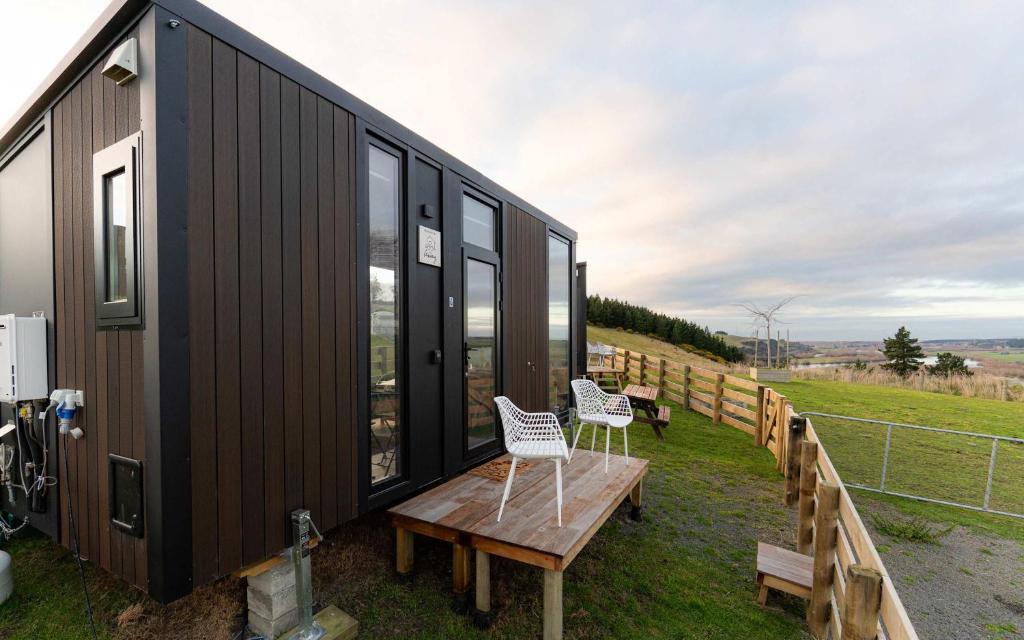 a black tiny house with two chairs and a bench at Bruce Farm Alpaca Cabin 4 by Tiny Away in Balclutha