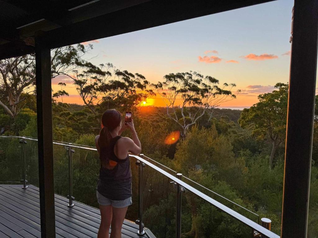 Bushys Hilltop (Kingfisher Bay, Kgari) في فريزر آيلاند: a woman taking a picture of the sunset from a balcony