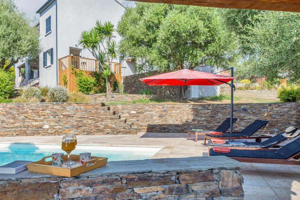 una piscina con un ombrellone rosso e sedie e un tavolo con un bicchiere di vino di Villa Le Corps de Garde a Furiani