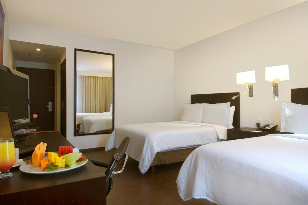 A modern two-bed room includes white bedding, wall lights, and a small dining or work table.