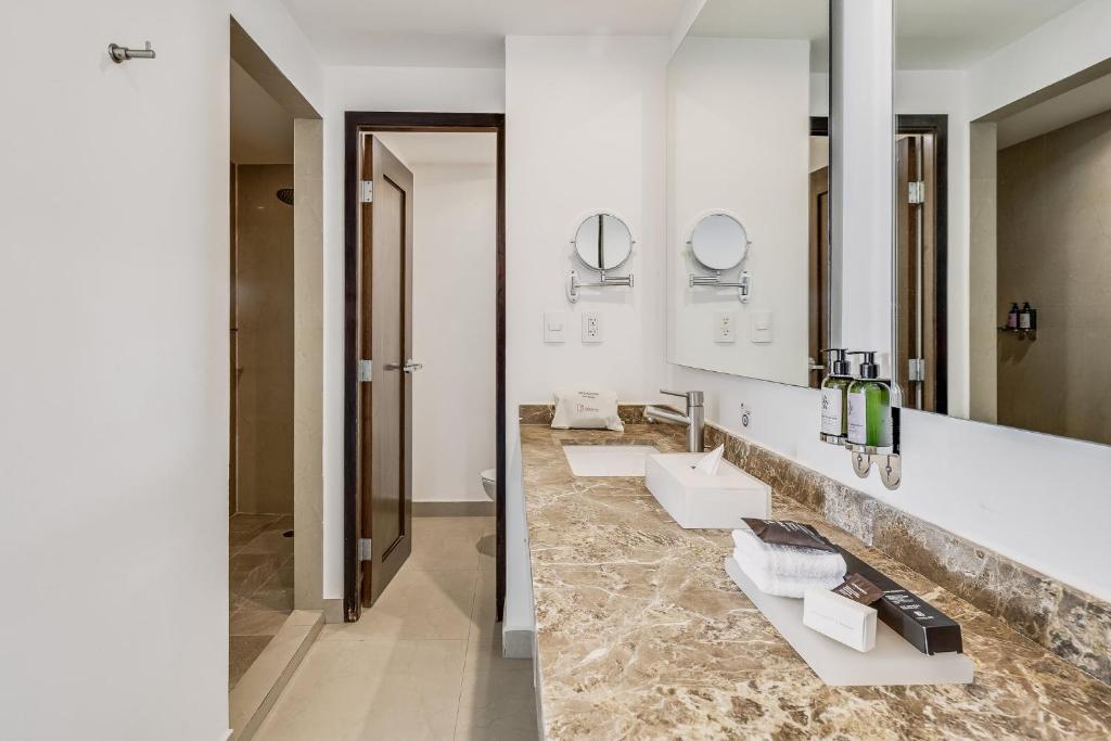A bright bathroom vanity has two sinks, a long mirror, and a stone-look counter.