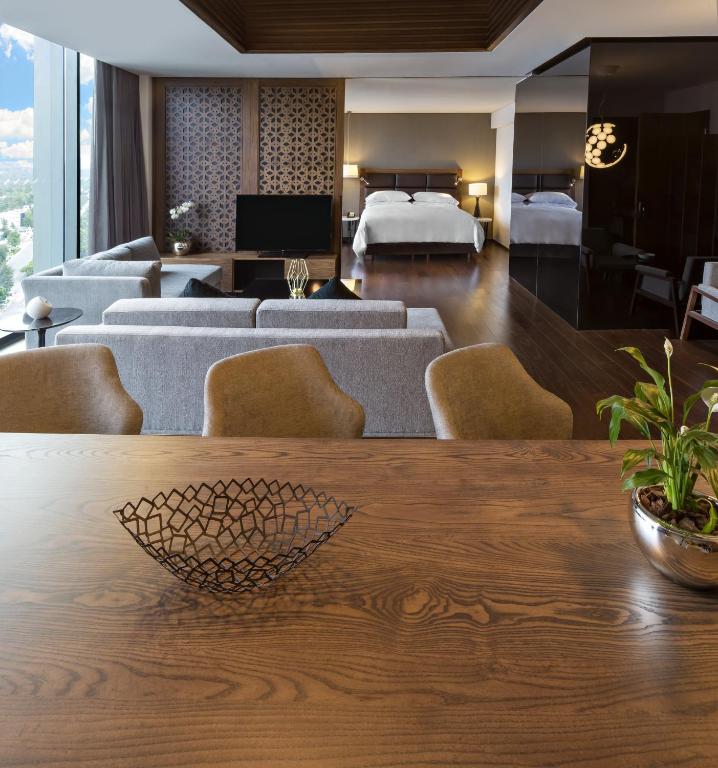 A high-rise suite presents a king bed, lounge seating and a dining table against a bright city backdrop.