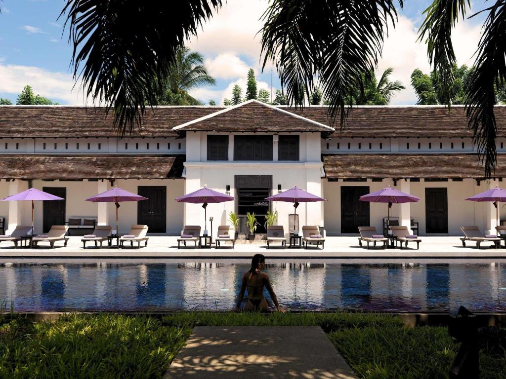 a woman sitting next to a swimming pool with purple umbrellas at Sofitel Luang Prabang in Luang Prabang