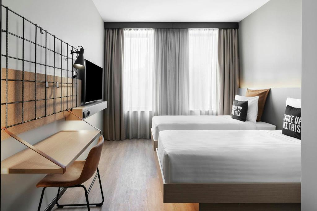 A twin room includes a wall-mounted television, a narrow desk, and tall curtained windows.