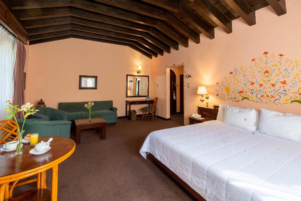 A large traditional room combining a bed, two sofas, and a small dining table beneath exposed beams.