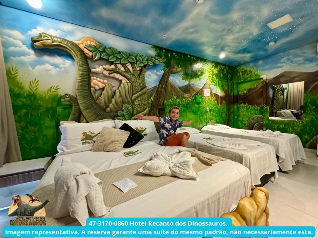 a man sitting on a bed in a room with a dinosaur mural at Hotel Recanto dos Dinossauros in Penha