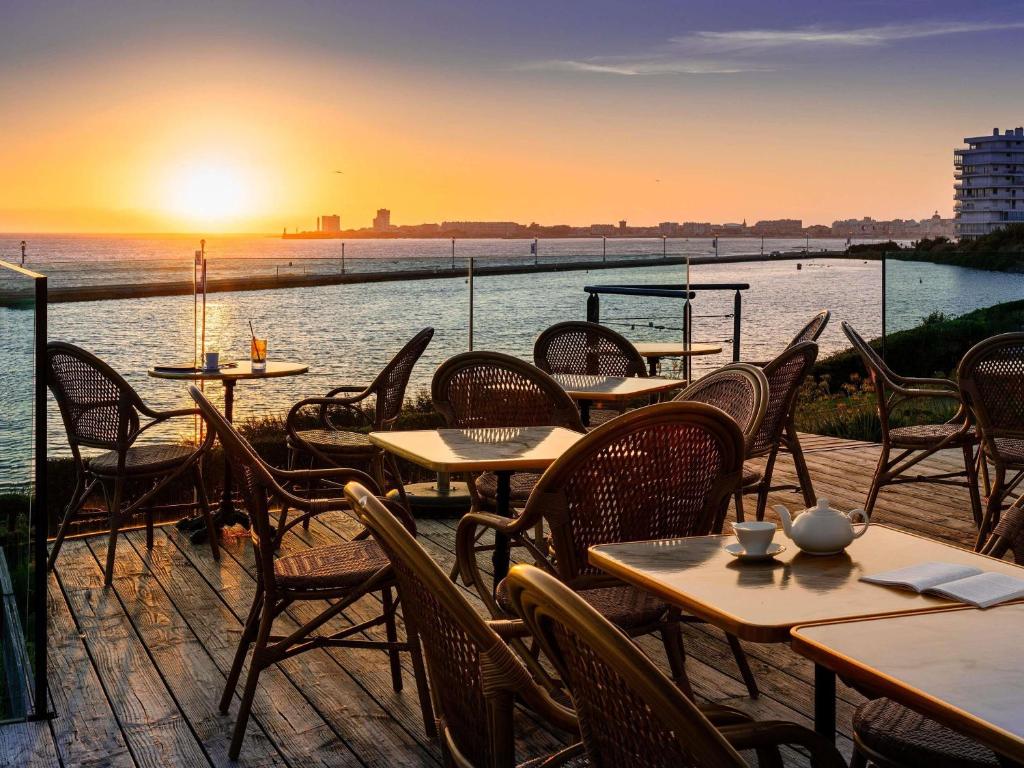 eine Terrasse mit Tischen und Stühlen und das Meer bei Sonnenuntergang in der Unterkunft Côte Ouest Thalasso & Spa Les Sables d'Olonne - MGallery Collection in Les Sables-dʼOlonne
