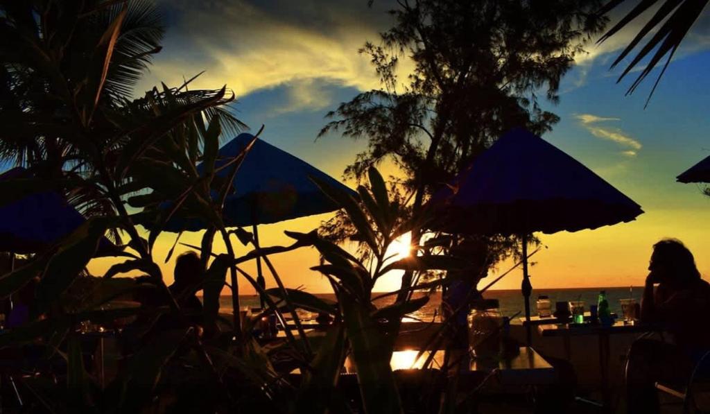une femme assise à une table sous des parasols à la plage dans l'établissement COTTAGE OCEAN classé 3 étoiles - 4 à 6 personnes - 2 chb - Accès direct à la plage et à l'esplanade de Boucan Canot avec ses bars et restaurants, à Saint-Gilles les Bains