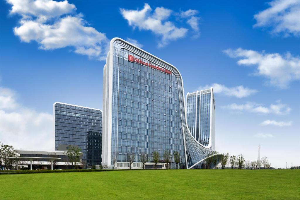 a large glass building with a green field in front of it at Hilton Garden Inn Zhengzhou Airport Aerotropolis in Zhengzhou