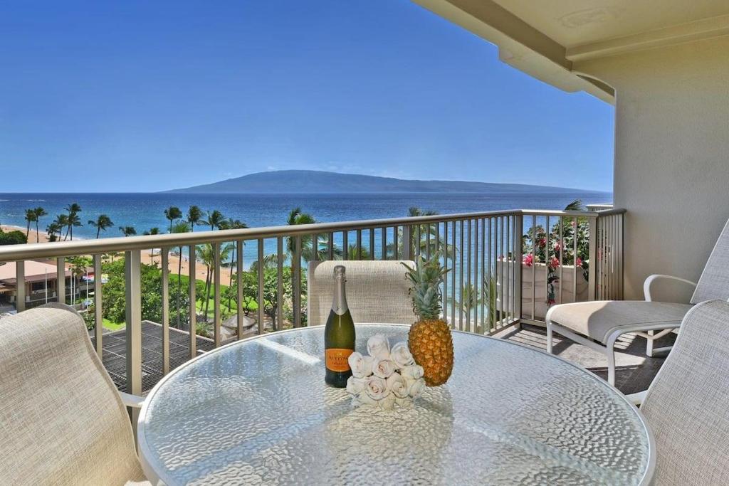 a glass table with a bottle of wine and pineapple on a balcony at Whaler 859 BeachFront Luxury Condo w Ocean Views in Lahaina