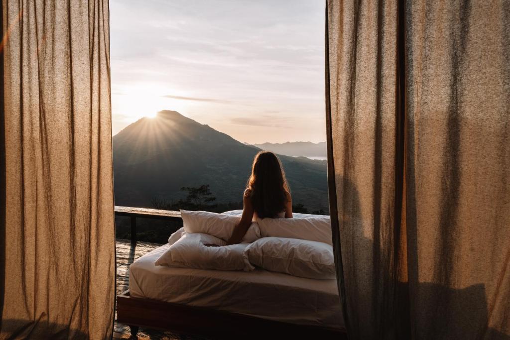 a woman sitting on a bed looking out a window at Batur Cabins in Kintamani