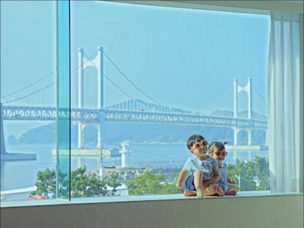 two children sitting on a window sill with a view of a bridge at 광안사계스테이 GwanganSagyeStay in Busan