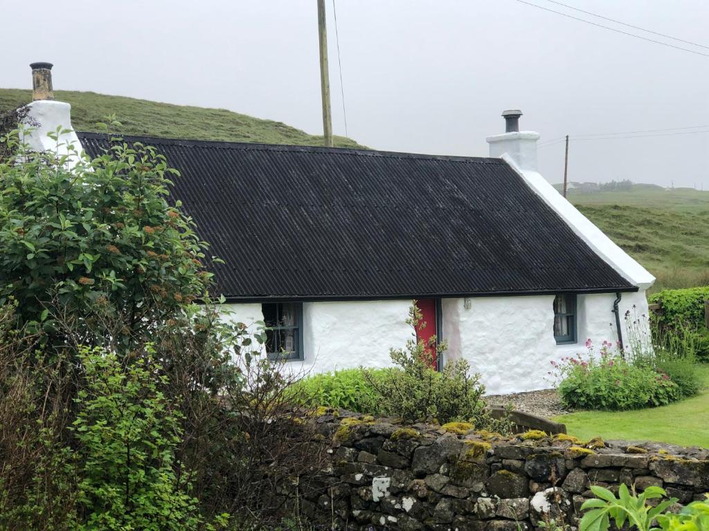 a white house with a black roof and a stone wall at Allt Mor Cottage in Milovaig