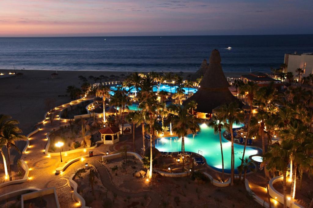 Aerial view of illuminated pools, beach, palm trees, and ocean at dusk.
