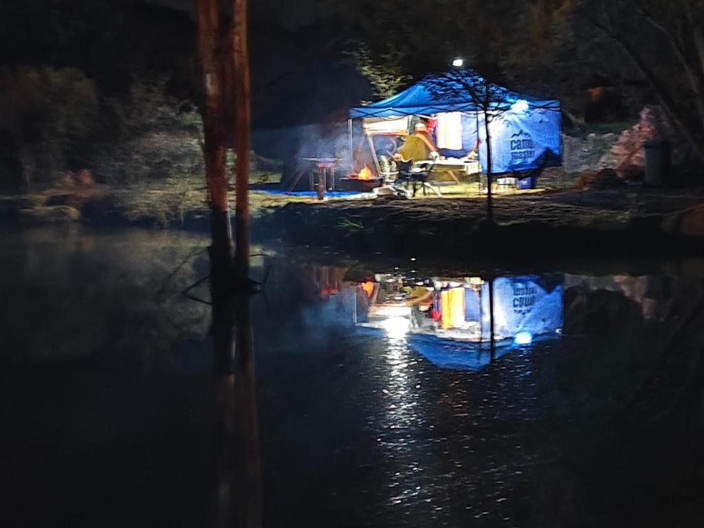 Un hombre sentado en una tienda de campaña junto a un río por la noche en CradleLicious camp & caravan, en Beckedan