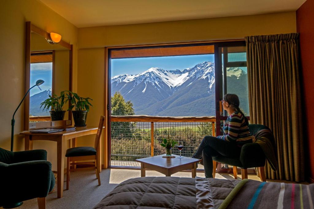 a woman sitting in a chair looking out a window at a mountain at Wilderness Lodge Arthurs Pass in Arthur's Pass