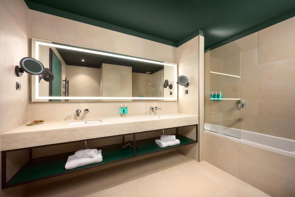 Double-sink bathroom with backlit mirror, wall-mounted magnifying mirror, bathtub, towel shelf and green ceiling at Novotel Zagreb in Zagreb