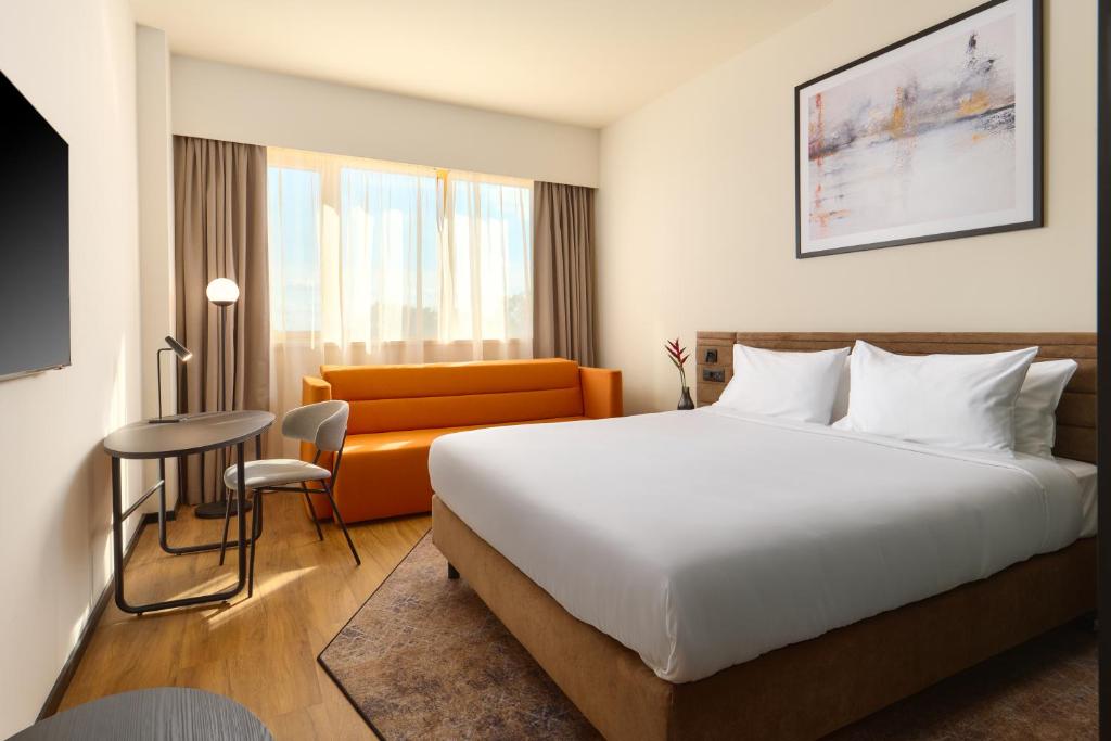 King bed with white linens, wall-mounted TV, work desk and lamp, orange sofa and floor-to-ceiling window with city view at Novotel Zagreb in Zagreb
