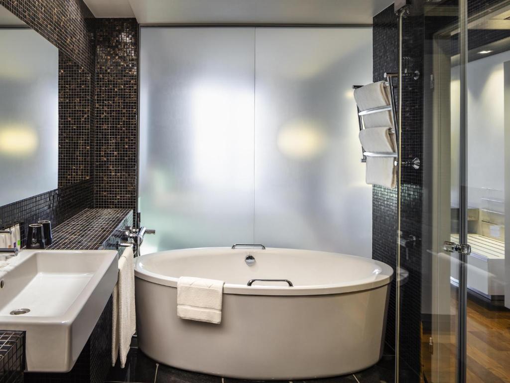 Freestanding oval tub with towel, wall-mounted sink, frosted glass shower and heated towel rack against black mosaic tile at Swissotel Tallinn in Tallinn