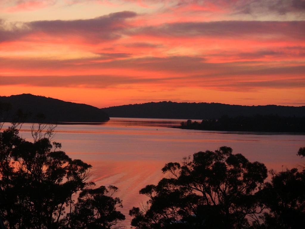 a sunset over a lake with trees in the foreground at A LAKEHOUSE ESCAPE - a Waterfront Reserve on shores of Lake Macquarie in Bonnells Bay
