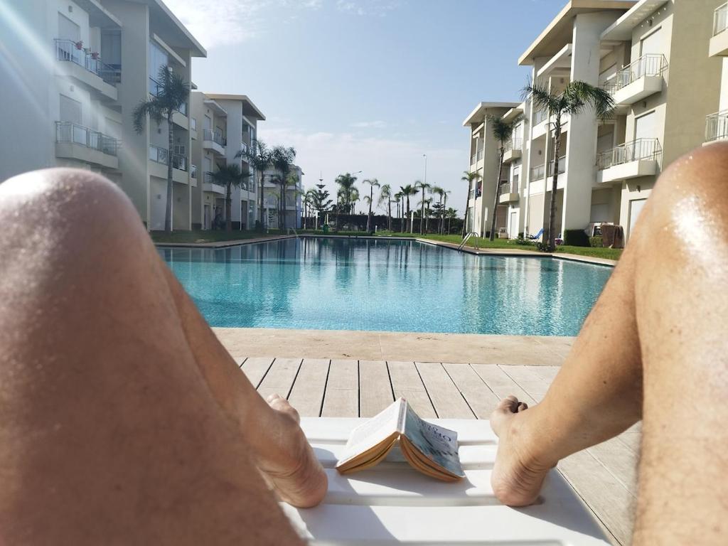 a man sitting on a towel reading a book next to a swimming pool at Apartment in residence CASABAY in Sidi Rahal