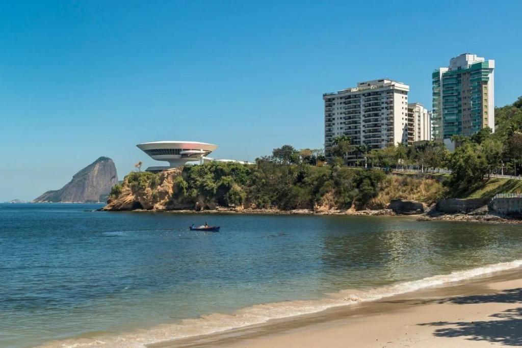 a beach with a boat in the water and buildings at Apartamento Inteiro Novo Praia, 2 Varandas, Piscina e Vista Parcial MAC in Niterói