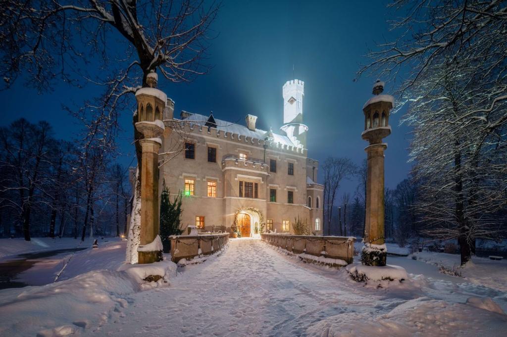 Snow-covered castle at night, illuminated entrance and stone bridge flanked by ornate pillars.