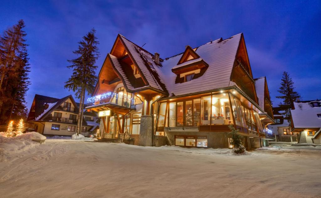 Alpine chalet at dusk with snow-covered roof, warm interior lights, festive string lights and tall pine trees.