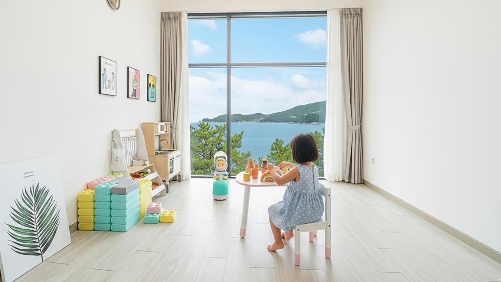 Una niña sentada en una mesa en una habitación con ventana. en Yeosu Sea&Blue Family Pension, en Yeosu