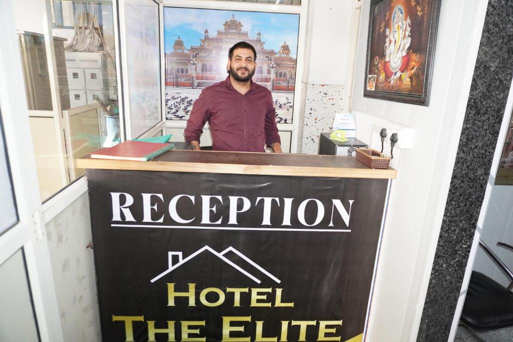 un hombre parado detrás de un mostrador con una señal en Hotel The Elite, en Jaipur