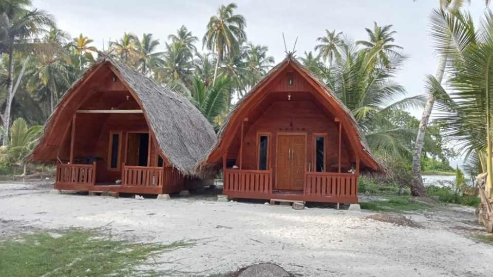 two thatched cottages on a beach with palm trees at North Pagai Bungalows in Pulau Silabusabeu