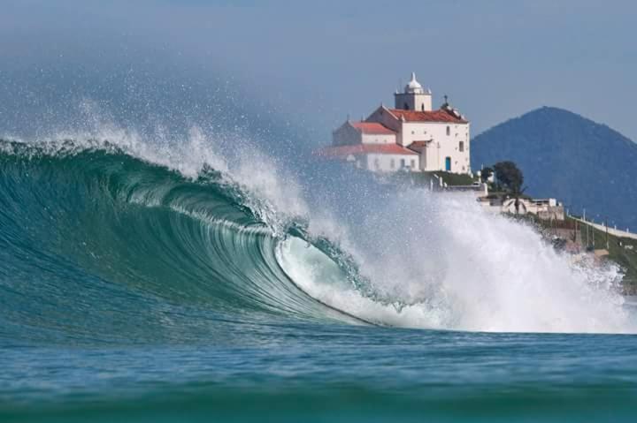 a wave in the ocean with a lighthouse in the background at WSL Saqua, Suite com ar, piscina, lounge, salão festas, churrasqueira, jogos, jardim, estacionamento 800m2 in Saquarema