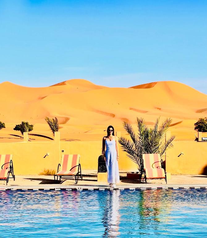 a woman standing next to a pool in front of a desert at Hotel Camping Les Roses Des Dunes & Piscine in Merzouga