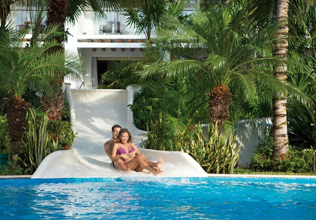 a man and a woman sitting on a white slide by a swimming pool at Secrets Aura Cozumel - Adults Only in Cozumel