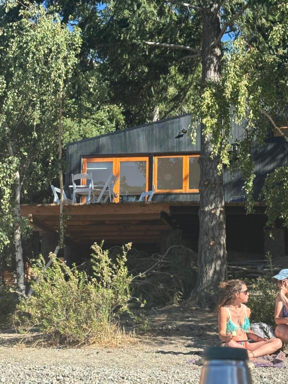two women sitting under a tree in front of a house at Cabaña Sobre Playa Lago Gutiérrez 2 in San Carlos de Bariloche