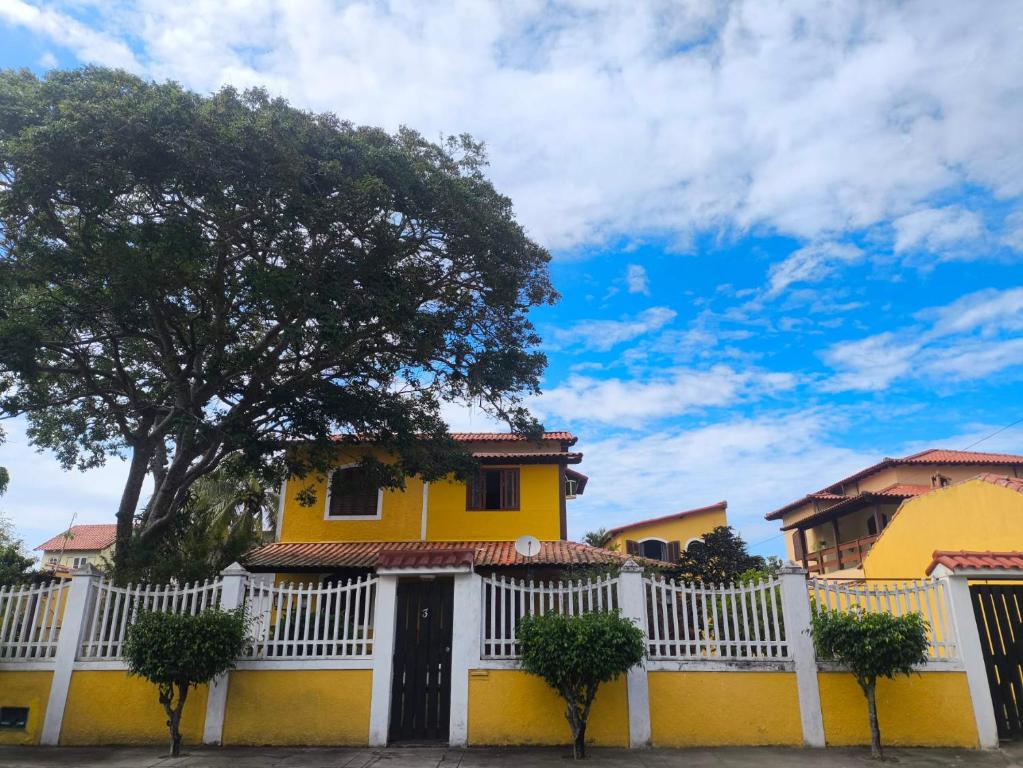 a yellow house with a white fence and a tree at Casinha Guest House in Saquarema