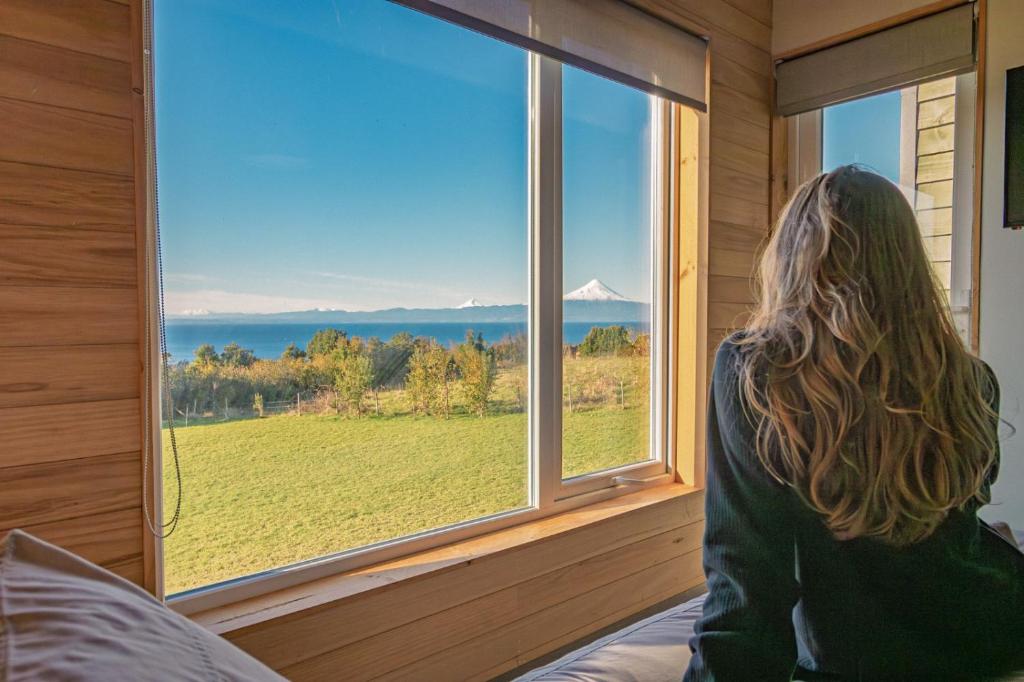 a woman sitting on a bed looking out a window at Ave Lodge Frutillar in Frutillar