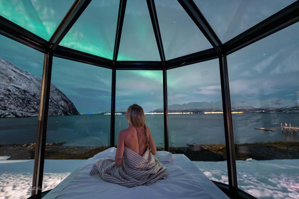 a woman sitting on a bed looking out at the water at Lyngen Aurora Resort in Koppangen