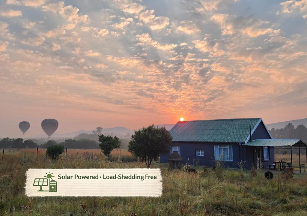 a house in a field with a sunset and hot air balloons at Firefinch Country Cottage in Brits