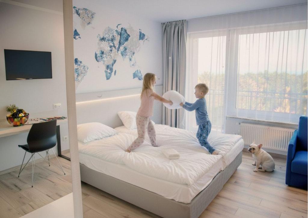 Two children playfully pillow-fighting on bed while small dog watches beside bright window.
